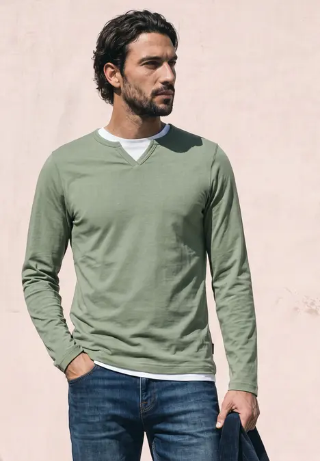 Een man met groen shirt met lange mouwen en spijkerbroek staat voor een lichte muur en kijkt opzij.
