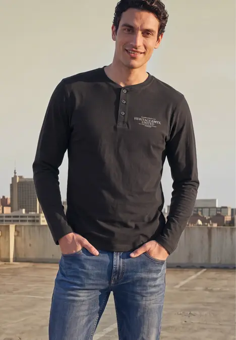 Un homme en t-shirt à manches longues sombre et jean bleu sourit sur un toit urbain.