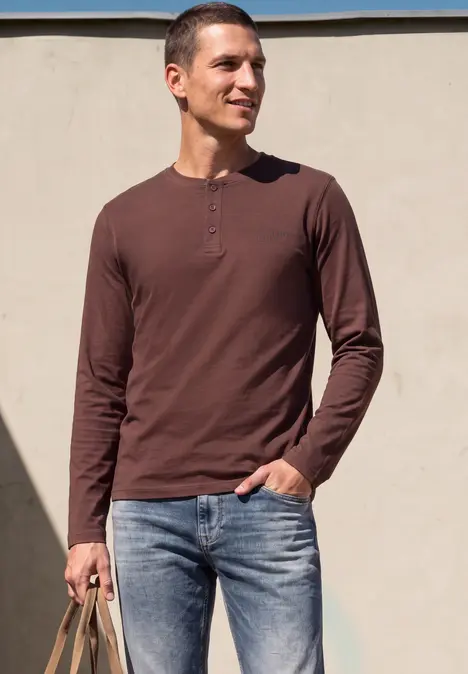 Homme en sweat marron à manches longues et jeans, tenant un sac tressé, debout devant un mur clair.