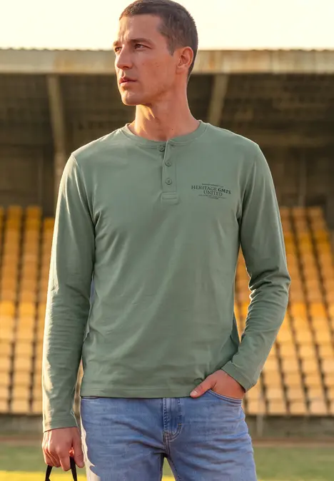 Un homme en t-shirt vert et jeans se tient dans un stade, tenant un smartphone dans la main.