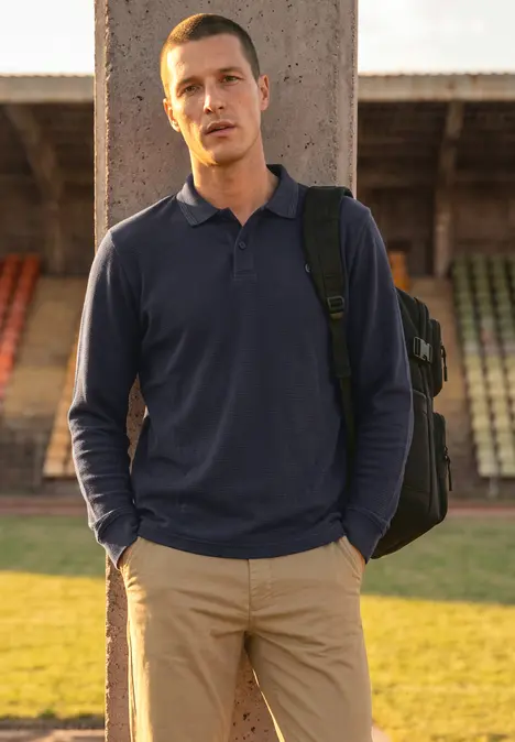 Ein Mann in blauem Poloshirt und beigen Hosen steht mit Rucksack vor leeren Tribünen.