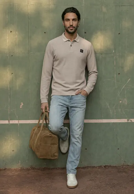 Ein Mann in beigem Polo, Jeans und weißen Sneakers lehnt an einer grünen Wand und hält eine braune Tasche.