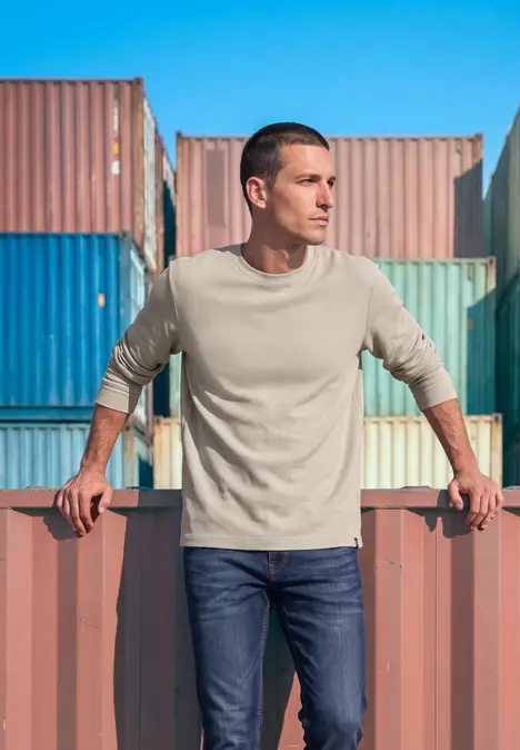 Ein Mann lehnt an bunten Frachtcontainern im Hafen, trägt beige Pullover und Jeans.
