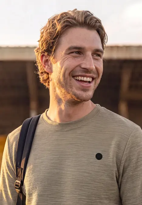 Ein junger Mann in einem beigen T-Shirt mit Rucksackschultergurt lächelt im Freien.