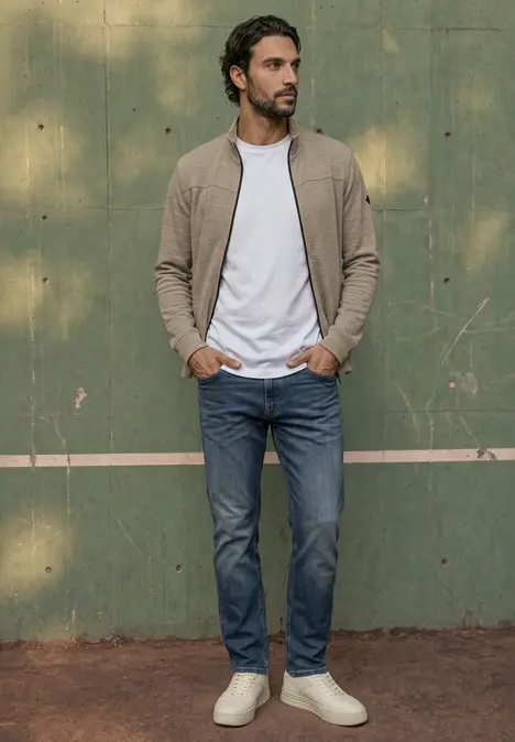 Ein Mann in Jeans, weißem Shirt und beiger Strickjacke steht vor einer grünen Wand und lehnt lässig.