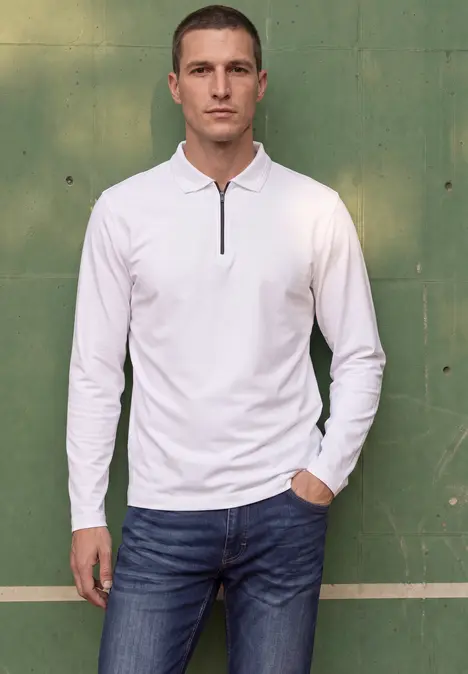 Ein Mann mit kurzem Haar in weißem Langarm-Polo und blauen Jeans steht vor grünem Hintergrund.
