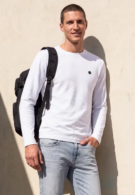 Ein junger Mann in weißem Langarmshirt, Jeans und Rucksack steht lässig vor einer Wand.