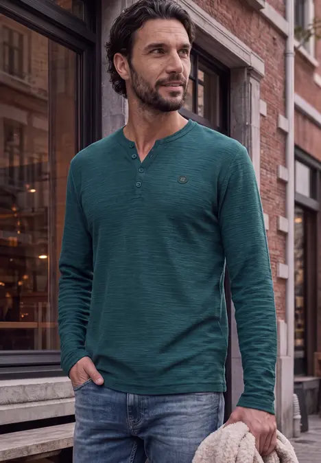 Een man draagt een groen shirt met lange mouwen en jeans en staat op een stadsstraat.