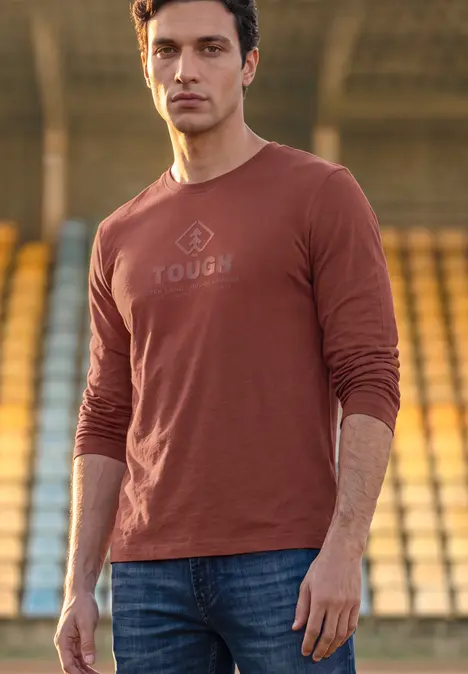 Mann in braunem Langarmshirt mit Aufschrift "TOUGH" vor Tribünen.