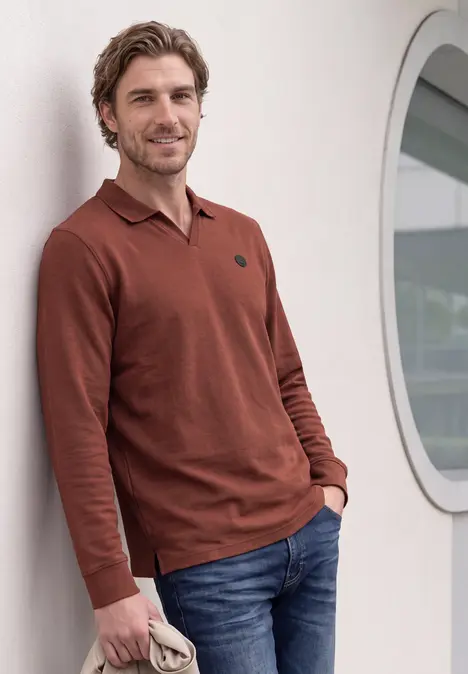 Ein Mann lehnt lässig an einer Wand, trägt einen kastanienbraunen Pullover und Jeans.