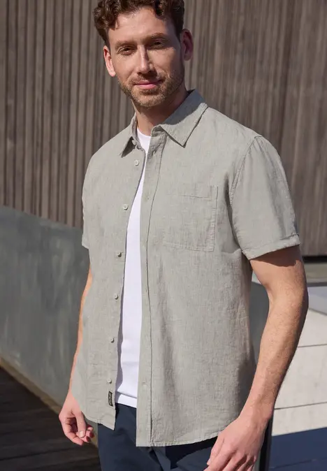 Ein Mann steht vor einer Holzfassade, trägt ein helles Hemd über einem weißen T-Shirt.