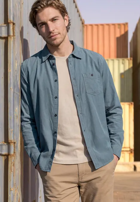 Ein junger Mann lehnt lässig an einem Container, trägt helles T-Shirt, offene Overshirt-Jacke und beige Hose.