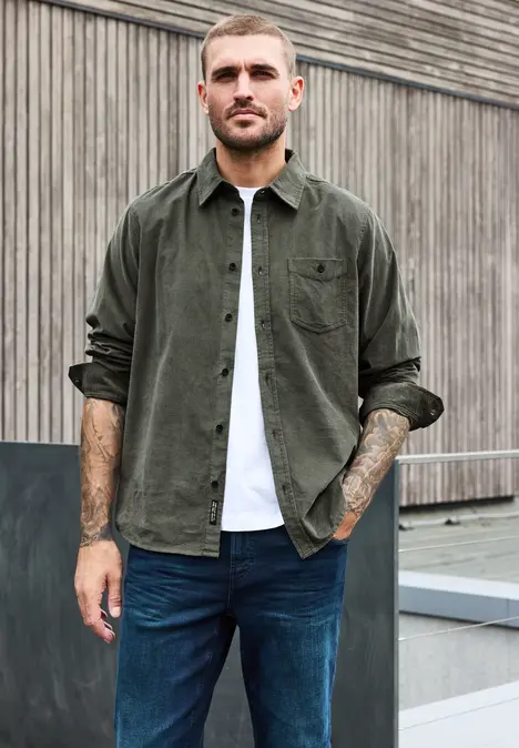 Ein tätowierter Mann in Jeans und Hemd posiert vor einer Holzfassade.
