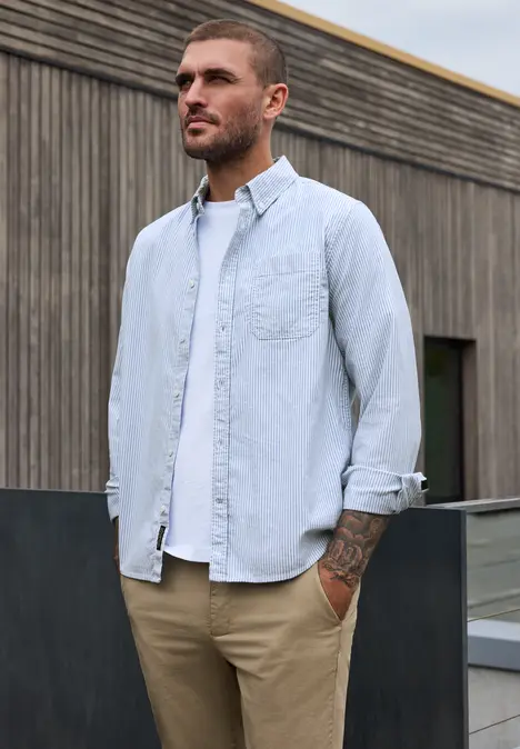 Ein Mann trägt hellblaues Hemd und beige Hose, steht vor einer Holzfassade und blickt in die Ferne.
