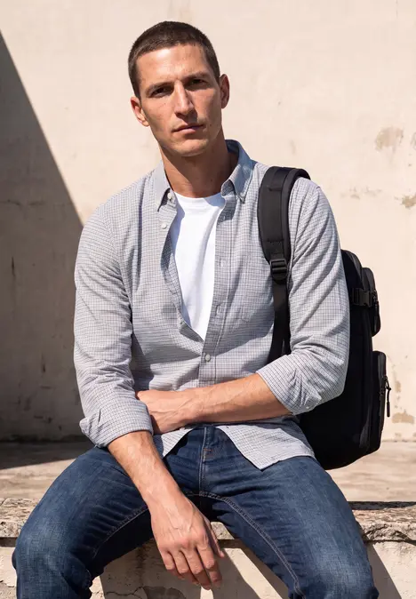 Ein junger Mann mit Rucksack sitzt locker in Jeans und Hemd vor einer hellen Wand.