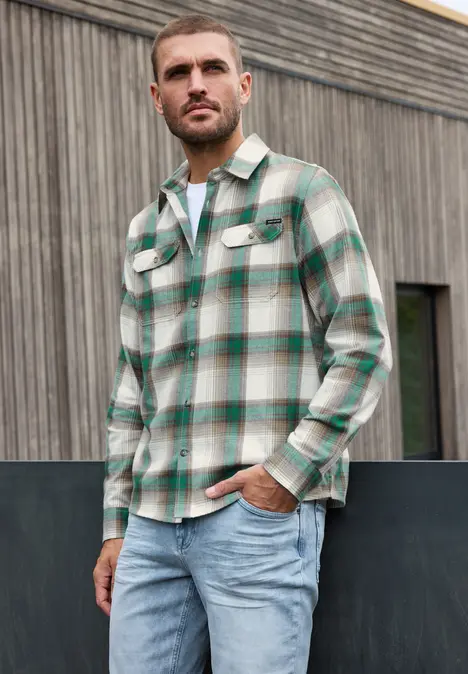 Ein Mann in kariertem Hemd und Jeans steht vor einer Holzfassade.