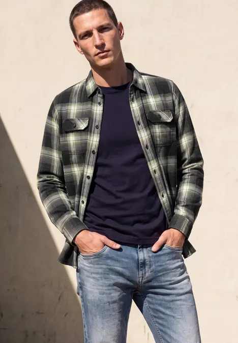 Ein junger Mann lehnt an einer Wand, trägt ein kariertes Hemd über einem dunklen T-Shirt und Jeans.
