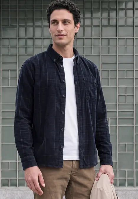 Ein junger Mann steht vor einer Glasfliesenwand, trägt weißes T-Shirt, offenes kariertes Hemd und braune Hose.