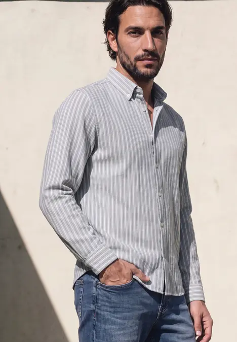 Un homme en chemise rayée et jean pose devant un mur clair, la main dans la poche.