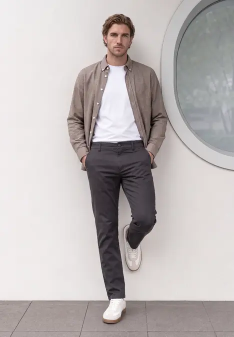 Ein Mann lehnt entspannt an einer Wand, trägt weißes T-Shirt, graue Hose, braune Jacke und weiße Sneaker.