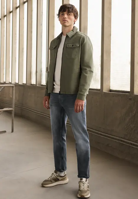 Ein Mann steht in lässiger Kleidung (grüne Jacke, weißes T-Shirt, Jeans, Sneaker) vor großen Fenstern.