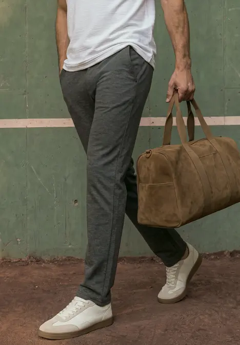 Un homme en pantalon gris et baskets blanches porte un sac de voyage marron sur fond vert.