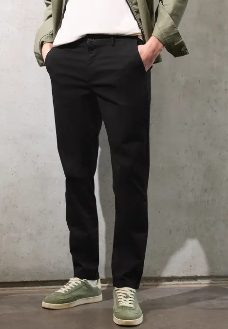 Czarne chinosy, zielone sneakersy, biały T-shirt i oliwkowa kurtka na tle betonu.