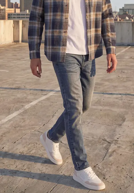 Ein Mann in Jeans, weißem T-Shirt, kariertem Hemd und weißen Sneakern auf einem Betonparkdeck.