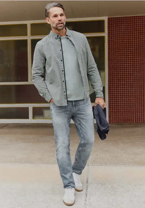 Ein Mann in lässigem Denim-Look mit Jeans, Hemd, T-Shirt und Sneakers hält eine Jacke vor einem Gebäude.