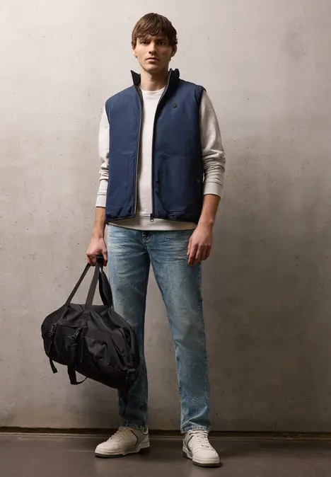Mann in blauer Weste, grauem Sweatshirt, Jeans und weißen Sneakern hält eine schwarze Reisetasche.