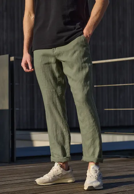 Ein schlanker Mann steht auf einem Holzdeck, trägt grüne Leinenhose, schwarzes T-Shirt und weiße Sneaker.