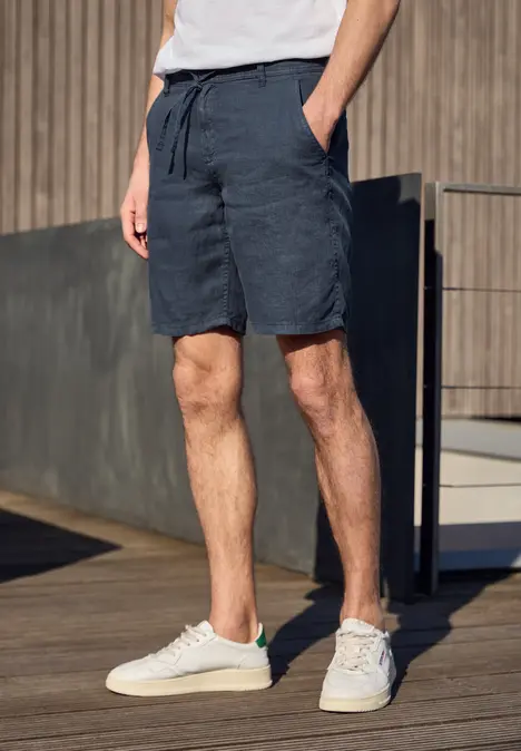 Mann steht auf Holzterrasse, trägt weiße Sneaker, dunkelblaue Shorts und weißes T-Shirt.