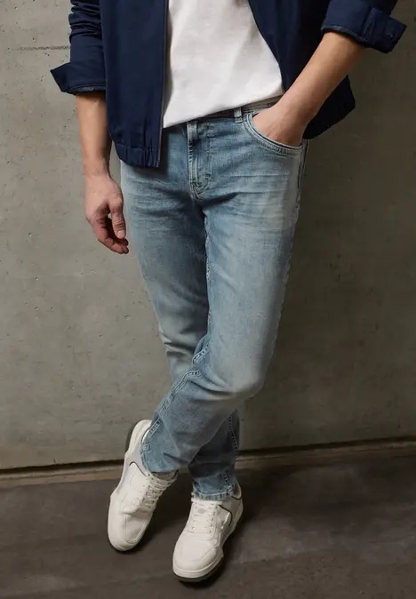 Person steht vor einer grauen Wand, trägt weiße Sneaker, blaue Jeans, weißes T-Shirt und blaue Jacke.