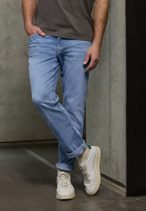 Ein Mann in hellblauen Jeans und weißen Sneakern lehnt lässig an einer Betonwand.