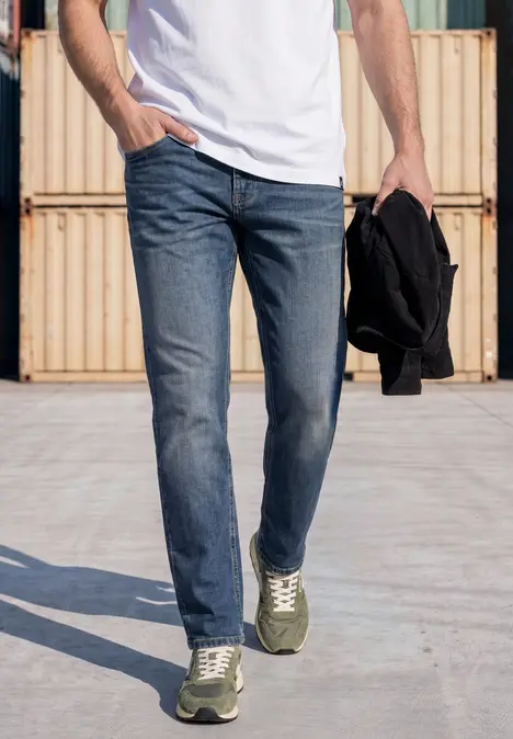 Mann in Jeans, weißem T-Shirt, grünen Sneakern, hält schwarze Jacke und geht an bunten Containern vorbei.