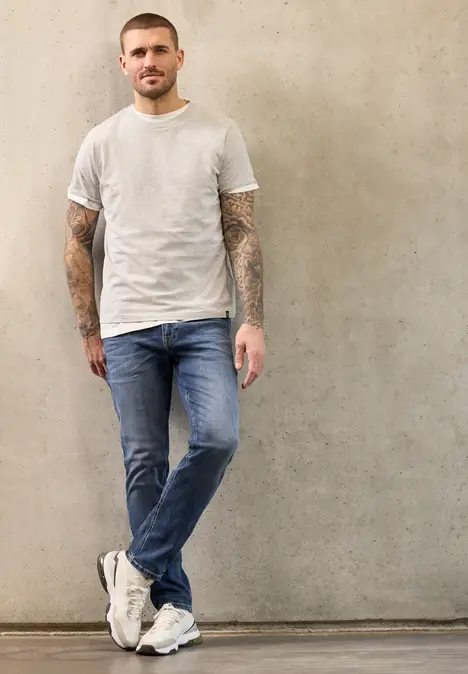 Ein tätowierter Mann lehnt in Jeans und weißem T-Shirt entspannt an einer grauen Wand.