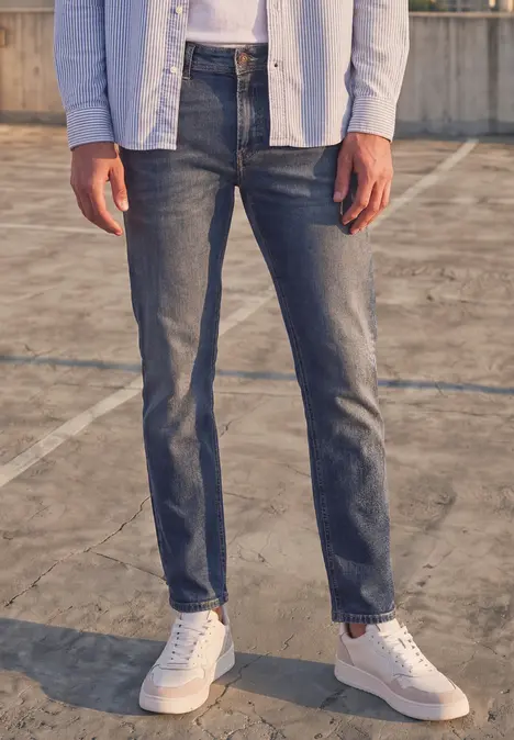 Mann in dunklen Jeans und weißen Turnschuhen steht auf einem Betonboden.
