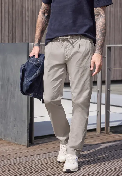 Ein Mann läuft auf Holzterrasse in hellen Chinohosen, weißen Sneakern, dunkelblauem T-Shirt und trägt eine Tasche.