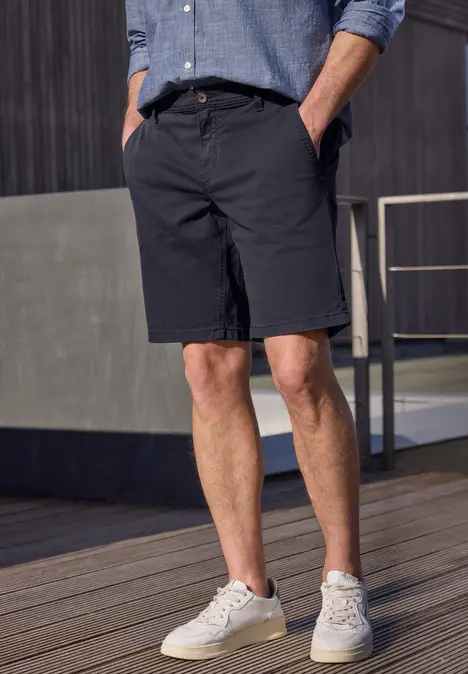 Homme en bermuda bleu marine, chemise bleu clair et baskets blanches, debout sur une terrasse en bois.