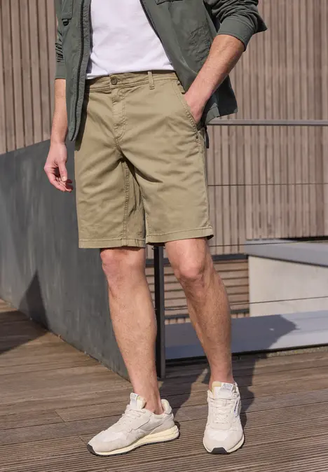 Een man in kaki shorts, wit shirt, groen jasje en lichte sneakers op een houten terras.