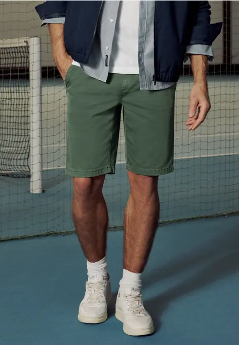 Mann steht vor einem Tennisnetz, trägt grüne Shorts, weiße Socken und Turnschuhe.