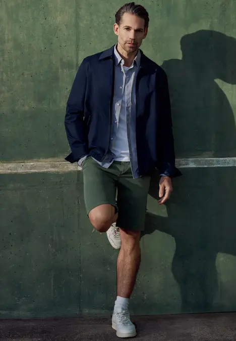 Homme en blazer bleu marine, chemise claire, shorts verts et baskets blanches adossé à un mur vert.