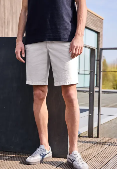 Een man met een zwart T-shirt, grijze bermuda en sneakers, staande op een houten terras voor een modern gebouw.