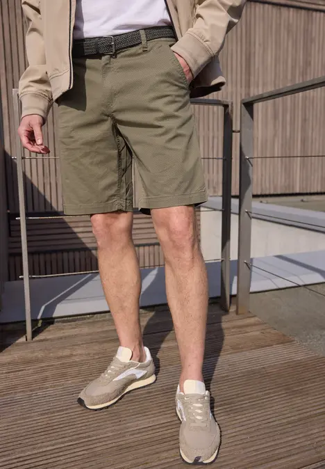 Un homme porte un short kaki, des baskets beiges et une veste légère sur une terrasse en bois.