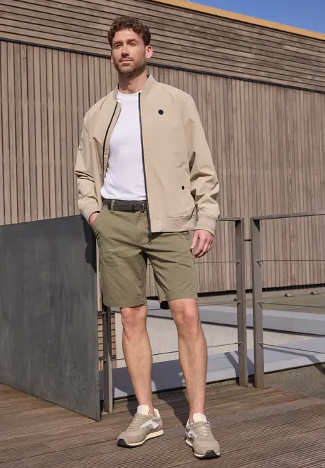 Ein Mann steht auf einer Holzterrasse, trägt beige Jacke, weißes Shirt, olivgrüne Shorts und Sneaker.