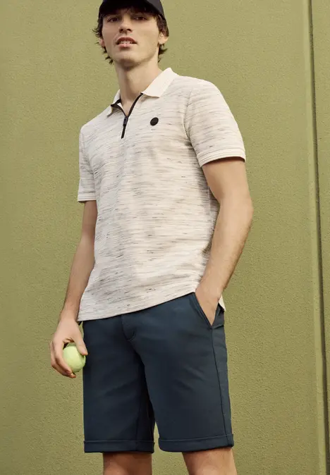 Mann in Poloshirt und Shorts hält einen Tennisball vor grüner Wand.