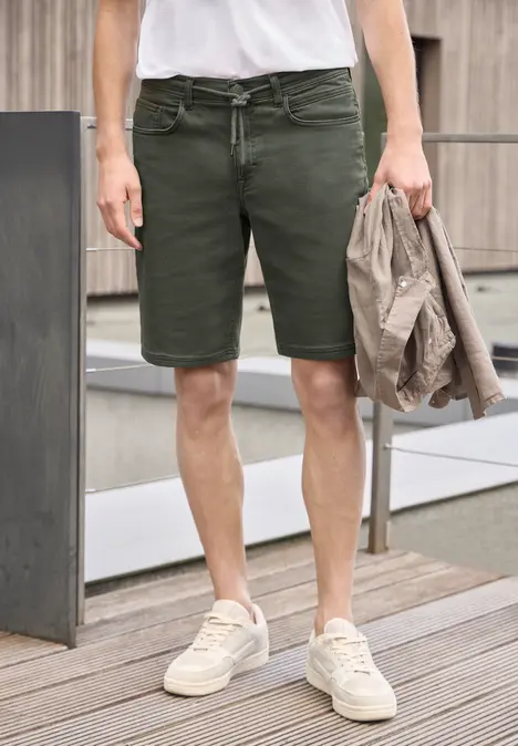 Een man draagt olijfgroene shorts en lichte sneakers, houdt een jas vast en staat op een houten terras.