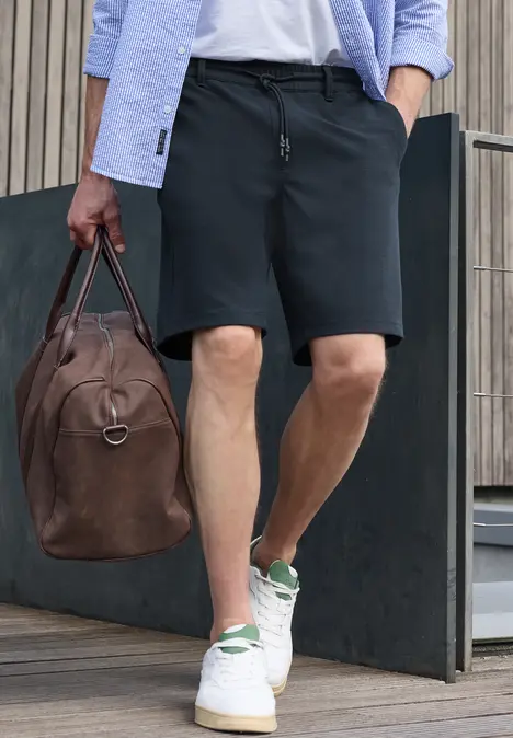 Ein Mann in schwarzen Shorts und weißen Sneakern hält eine braune Lederreisetasche und geht.