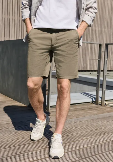 Ein Mann steht auf Holzdeck, trägt khakifarbene Shorts, weiße Sneaker und eine leichte Jacke.
