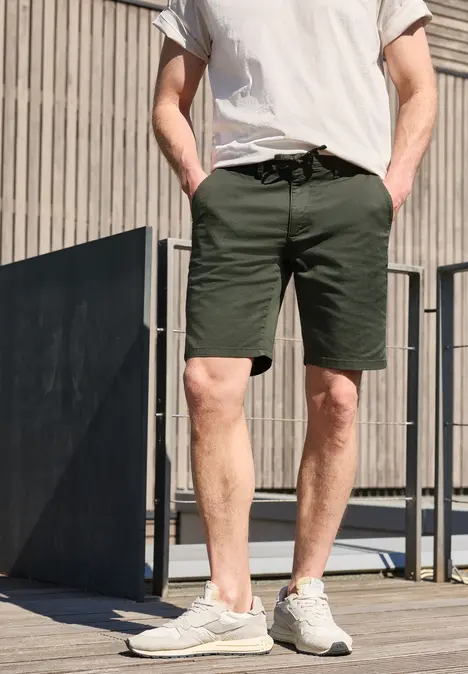 Een man in kaki short, wit t-shirt en sneakers, staand voor een houten gevel.